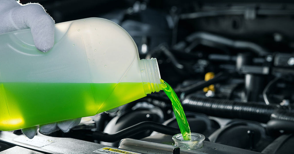 A close-up photo of a gloved hand holding a clear plastic jug of green antifreeze. The antifreeze is being poured into a car engine.